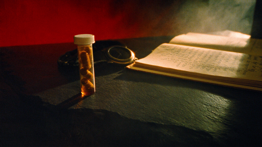 Cinematic editorial photograph — amber capsule vial on dark slate with leather journal and watch in shadow, golden-hour rim light. Brookhaven editorial style.